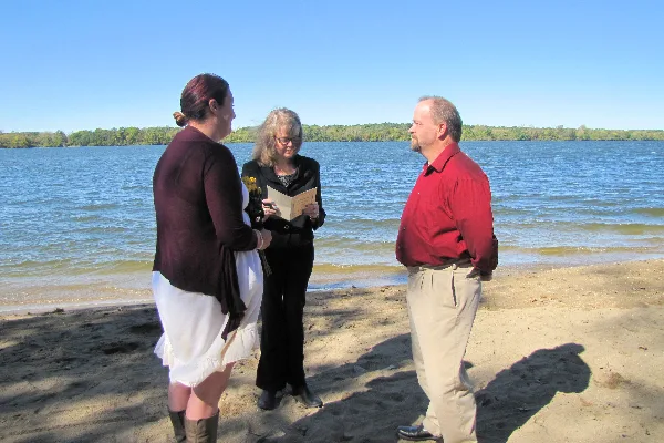 A wedding officiant reading from a book while standing between a bride and groom on a sandy beach.