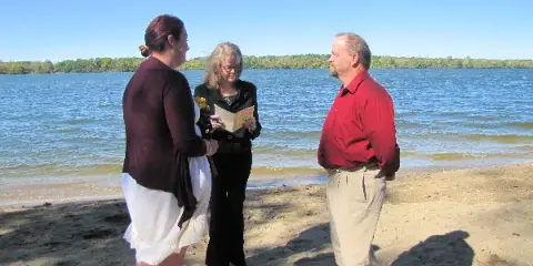 A wedding officiant reading from a book while standing between a bride and groom on a sandy beach.