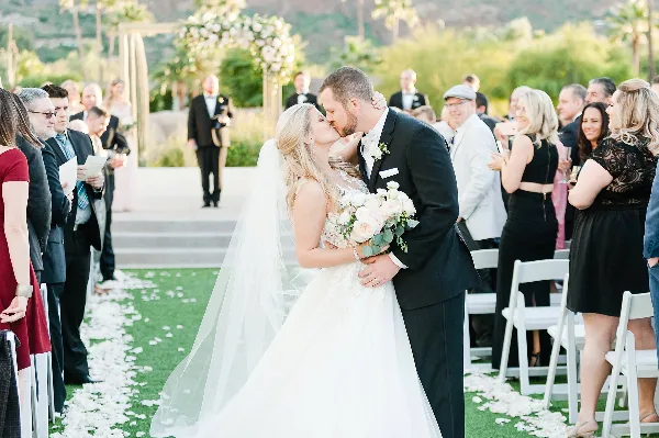 A bride and groom sharing a kiss at the altar during an outdoor wedding ceremony, surrounded by seated guests and floral decorations.