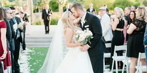 A bride and groom sharing a kiss at the altar during an outdoor wedding ceremony, surrounded by seated guests and floral decorations.