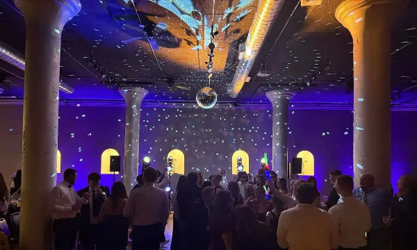 A crowded wedding dance floor with guests dancing under purple and blue stage lighting and a disco ball.