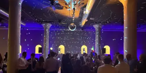 A crowded wedding dance floor with guests dancing under purple and blue stage lighting and a disco ball.