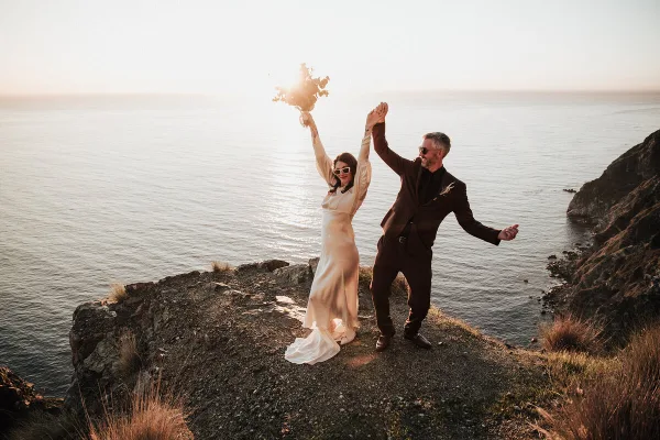 A bride and groom dancing on a rocky cliff overlooking the ocean at sunset.