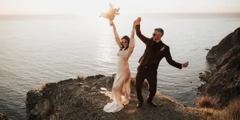 A bride and groom dancing on a rocky cliff overlooking the ocean at sunset.