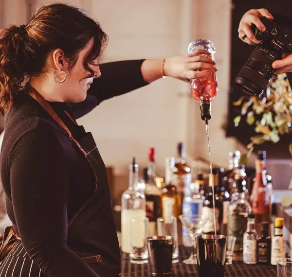 A bartender in an apron pouring a liquid from a bottle into a cocktail shaker at a bar station.