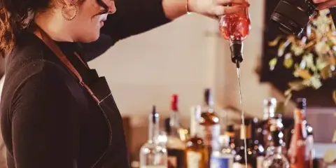 A bartender in an apron pouring a liquid from a bottle into a cocktail shaker at a bar station.
