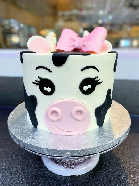 A custom cake decorated to look like a cow with a pink bow, sitting on a silver cake stand.
