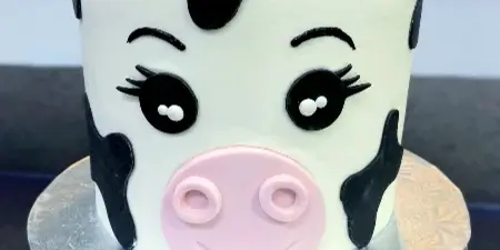 A custom cake decorated to look like a cow with a pink bow, sitting on a silver cake stand.