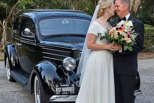 A bride and groom standing next to a vintage black car with a 'Just Married' license plate.