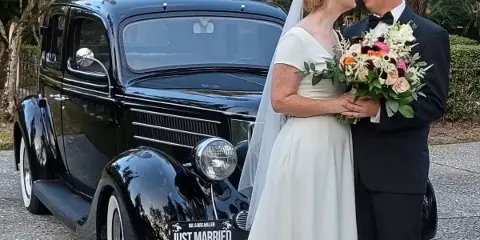 A bride and groom standing next to a vintage black car with a 'Just Married' license plate.