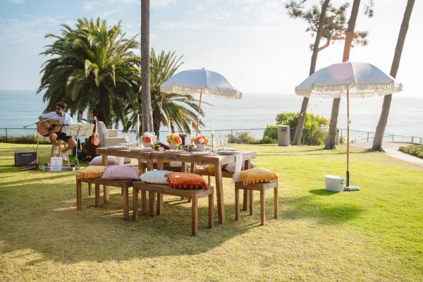 A professionally styled outdoor event table with a live musician playing guitar on a lawn near the ocean.