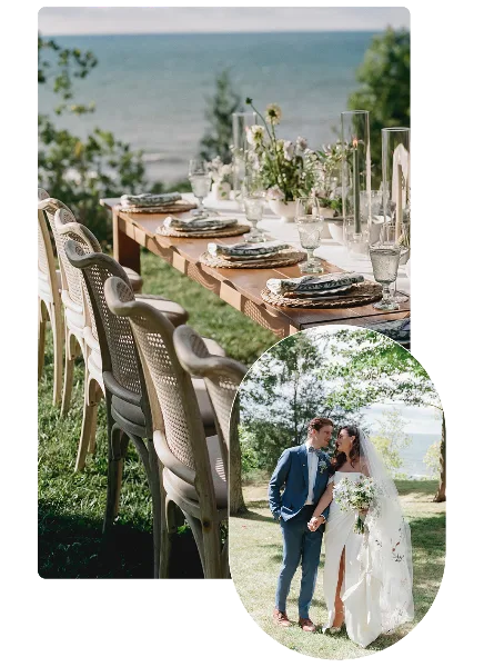 A wedding reception table set with floral centerpieces and glassware on a lawn near a body of water, with a bride and groom standing nearby.
