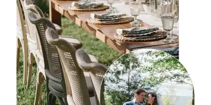 A wedding reception table set with floral centerpieces and glassware on a lawn near a body of water, with a bride and groom standing nearby.