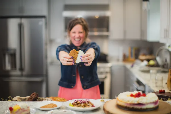 A smiling woman in a denim jacket holds a cookie toward the camera in a kitchen, with plated dishes and a cheesecake on the counter.