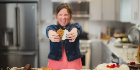 A smiling woman in a denim jacket holds a cookie toward the camera in a kitchen, with plated dishes and a cheesecake on the counter.