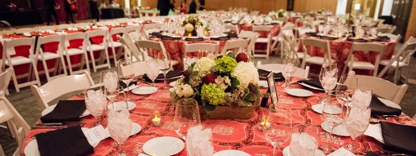 A wide shot of a formal event venue featuring round tables with red patterned tablecloths, white chairs, and elaborate floral centerpieces.