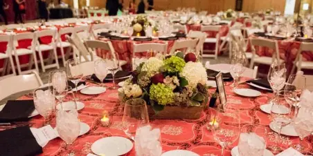 A wide shot of a formal event venue featuring round tables with red patterned tablecloths, white chairs, and elaborate floral centerpieces.