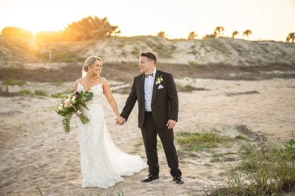 A bride in a white lace gown and a groom in a black suit holding hands on a sandy beach at sunset.