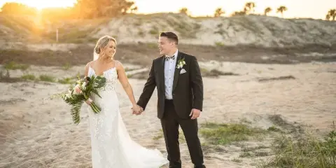 A bride in a white lace gown and a groom in a black suit holding hands on a sandy beach at sunset.