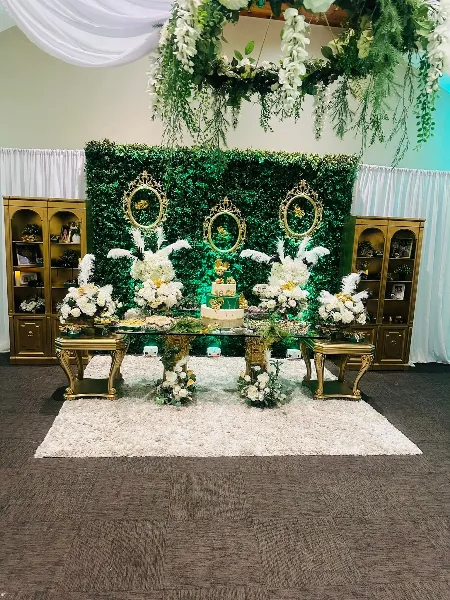 A professionally decorated event space featuring a greenery wall, gold display cabinets, and a dessert table with floral centerpieces.