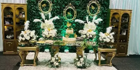 A professionally decorated event space featuring a greenery wall, gold display cabinets, and a dessert table with floral centerpieces.
