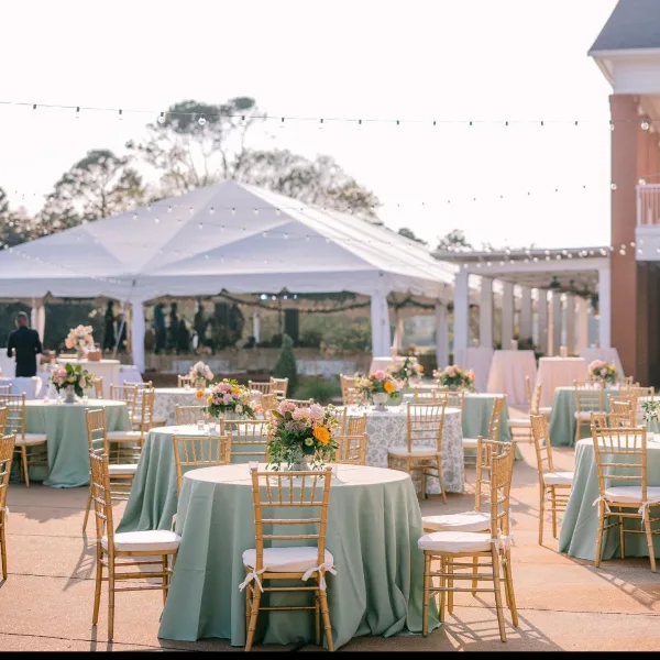 Outdoor wedding reception setup featuring a large white tent, string lights, and round tables with floral centerpieces.
