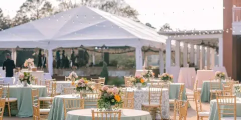 Outdoor wedding reception setup featuring a large white tent, string lights, and round tables with floral centerpieces.