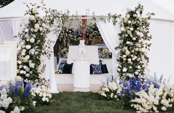 A wedding event setup featuring a white fluted pedestal, a white bench with pillows, and a white door frame decorated with flowers.