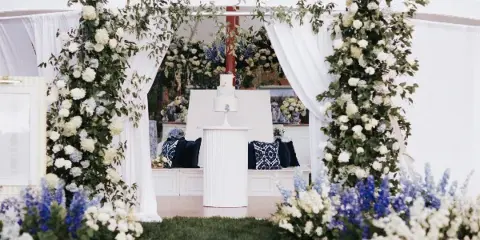 A wedding event setup featuring a white fluted pedestal, a white bench with pillows, and a white door frame decorated with flowers.