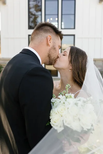 A bride and groom sharing a kiss outdoors, with the bride holding a bouquet of white roses.