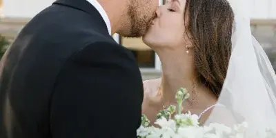 A bride and groom sharing a kiss outdoors, with the bride holding a bouquet of white roses.