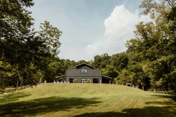 A rustic dark grey barn venue building on a grassy hill surrounded by trees and a blue sky.