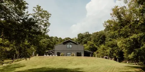 A rustic dark grey barn venue building on a grassy hill surrounded by trees and a blue sky.