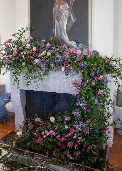 A lush, garden-style floral arrangement with pink, purple, and blue flowers and trailing greenery decorating a white fireplace mantel.