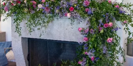 A lush, garden-style floral arrangement with pink, purple, and blue flowers and trailing greenery decorating a white fireplace mantel.