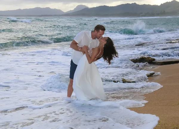 A bride and groom kissing on a sandy beach with ocean waves in the background.