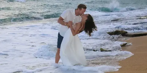 A bride and groom kissing on a sandy beach with ocean waves in the background.