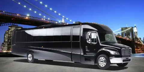 A black luxury charter bus parked in front of the Manhattan Bridge and New York City skyline at night.