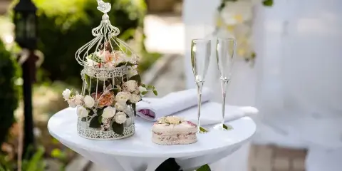 A white round table set with a tablecloth, two champagne flutes, and a decorative birdcage centerpiece filled with flowers.