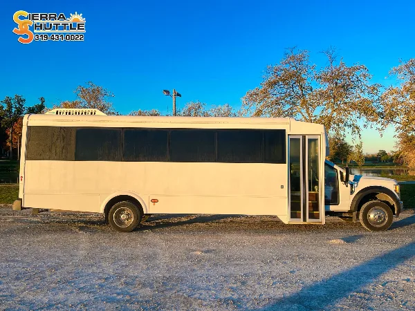A white shuttle bus parked on a gravel lot near a body of water with trees in the background.