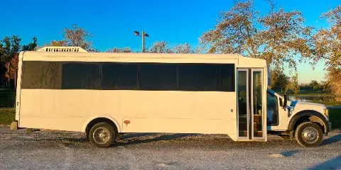 A white shuttle bus parked on a gravel lot near a body of water with trees in the background.