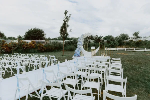 An outdoor wedding ceremony setup featuring white chairs, a floral arch, and a white aisle runner on a grassy lawn.