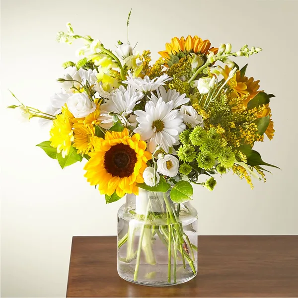 A mixed bouquet of white daisies, sunflowers, and greenery arranged in a clear glass vase on a wooden table.
