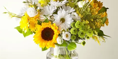 A mixed bouquet of white daisies, sunflowers, and greenery arranged in a clear glass vase on a wooden table.