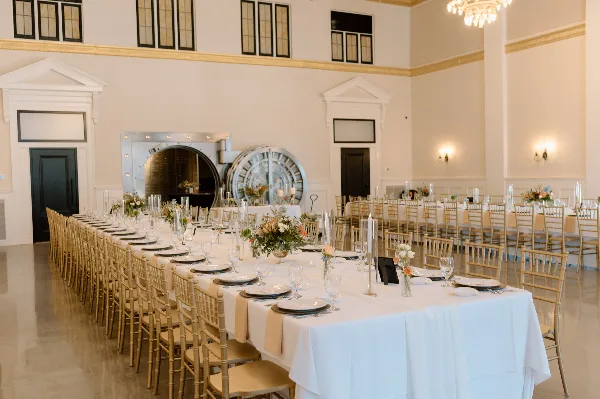 Interior view of a historic event venue ballroom featuring a large bank vault door, high ceilings, and a long banquet table set with white linens and floral centerpieces.