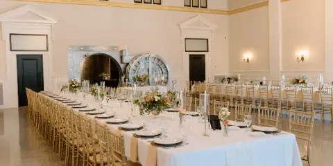 Interior view of a historic event venue ballroom featuring a large bank vault door, high ceilings, and a long banquet table set with white linens and floral centerpieces.