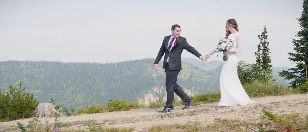 A bride and groom holding hands and walking on a scenic mountain overlook with a forested valley in the background.