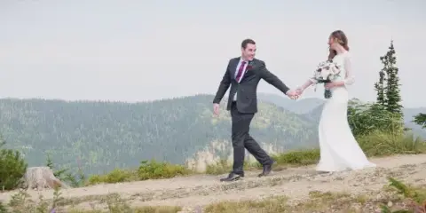 A bride and groom holding hands and walking on a scenic mountain overlook with a forested valley in the background.