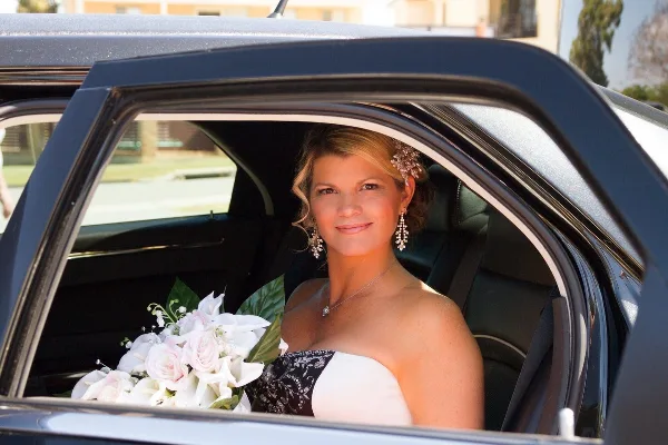 A bride in a wedding dress and holding a bouquet sits in the back seat of a black limousine.