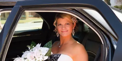 A bride in a wedding dress and holding a bouquet sits in the back seat of a black limousine.